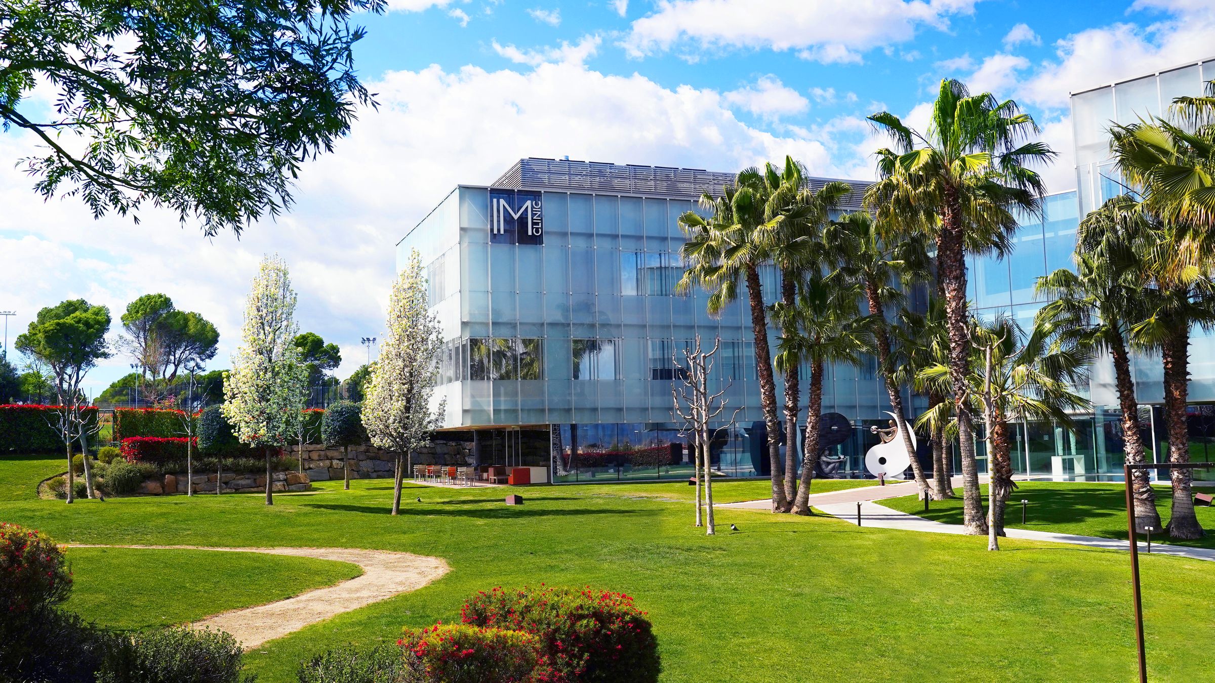 Fachada de cristal de IM Clinic entre palmeras, con el jardín delante